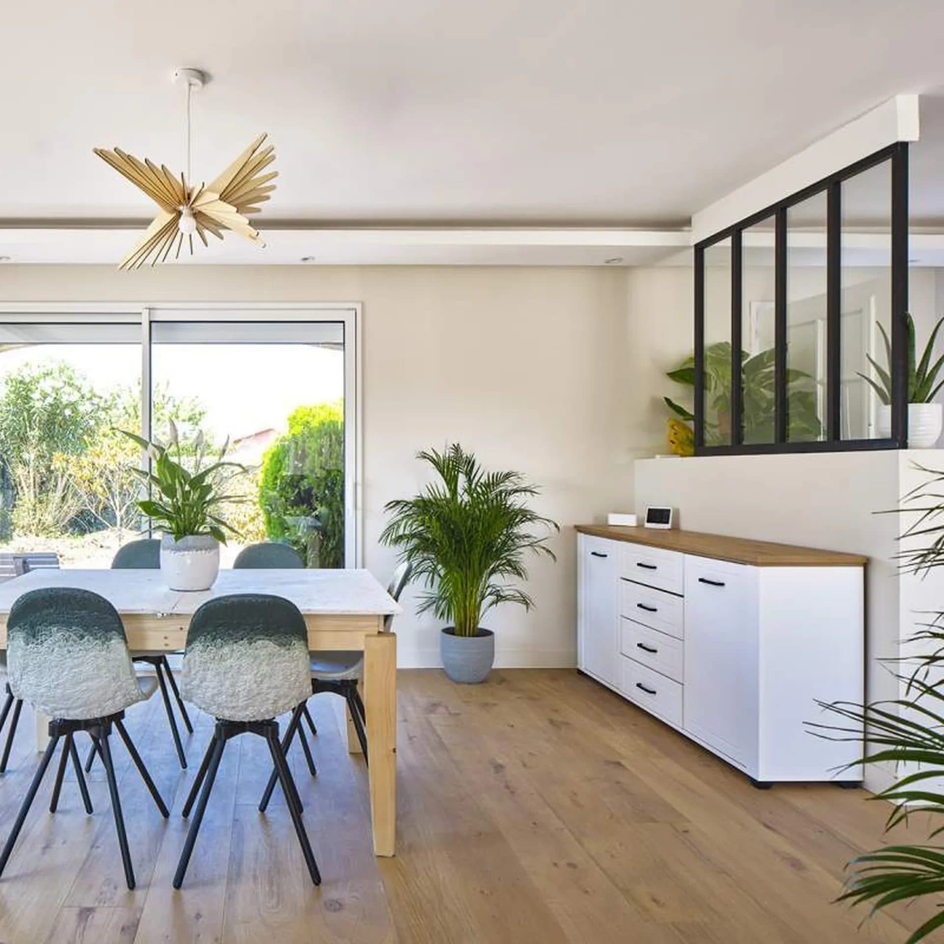 Salle à manger avec table en bois, suspension en laiton et cuisine blanche en second plan